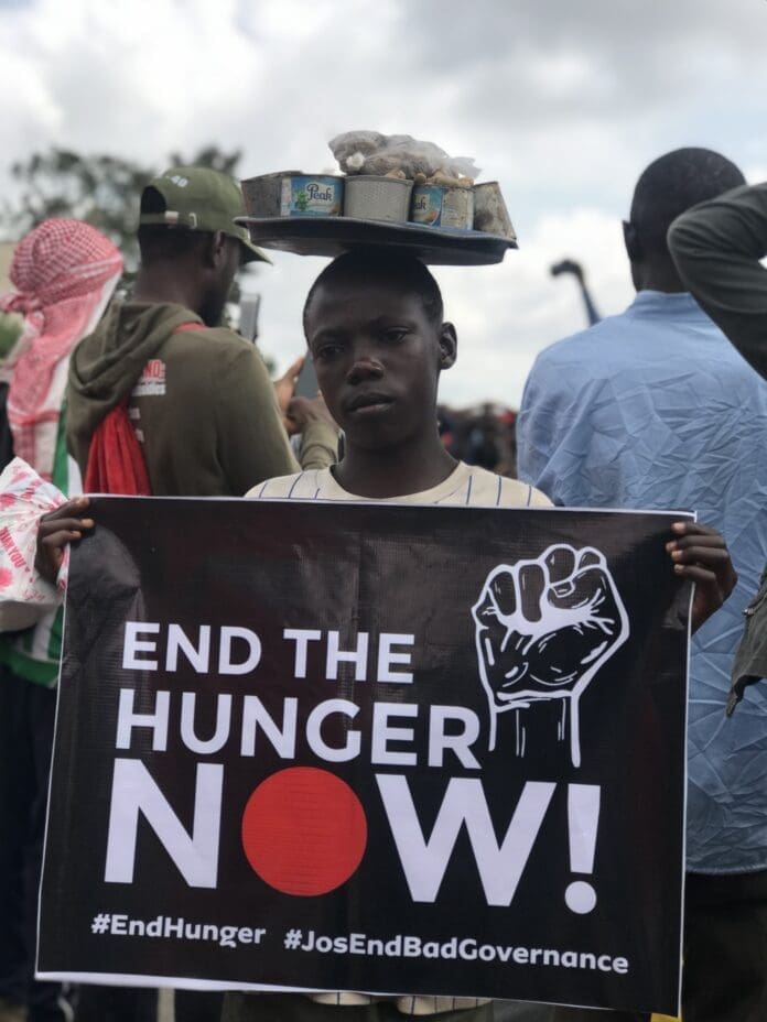 10year-old Abubakar Abdulhamid holds a placard while balancing a tray of peanuts on his head during #Endbadgovernance protest in Jos on August 4. Credit Masara Kim